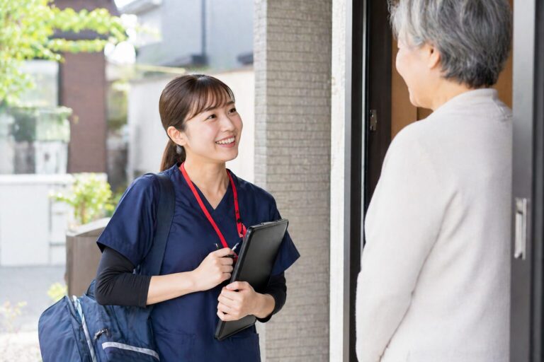 訪問先の玄関先で、バインダーを手に高齢の女性へ笑顔で挨拶する看護師