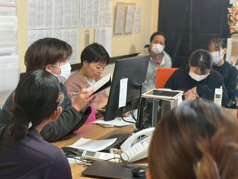 事務所で複数のスタッフがモニターを囲み、記録を見ながら話し合っている様子